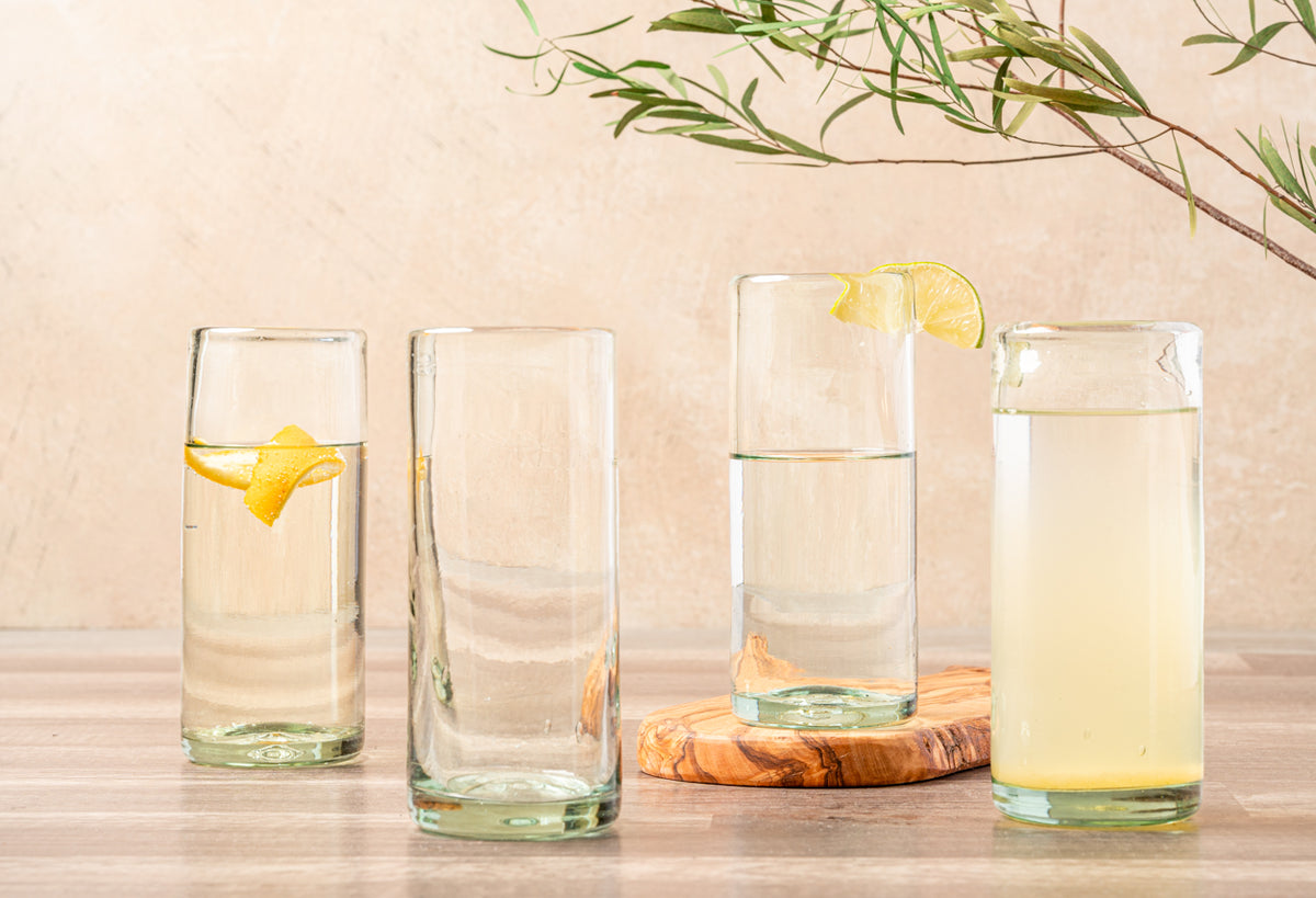 Four clear cylindrical glasses with varying liquid levels, one containing lemon slice, arranged on wooden surface.