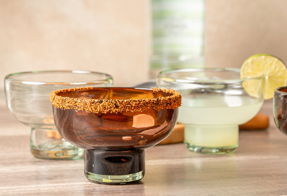 Two handblown glass margarita glasses, one with spiced rim and amber liquid, one with clear liquid and lime slice.