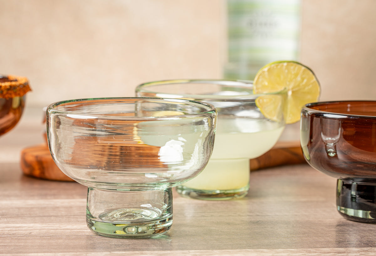 Two clear handblown glass margarita glasses with wide bowls and pedestal bases, one empty and one filled with pale yellow liq