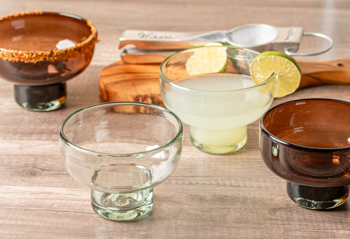 Two handblown margarita glasses, one clear and one with amber-colored rim, displayed with lime slices and cocktail.
