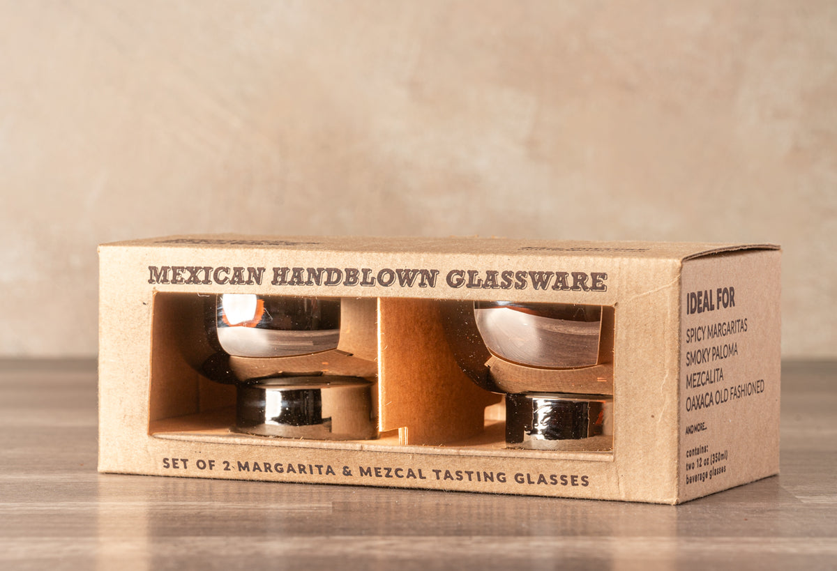 Two handblown margarita glasses in a cardboard box with wood-colored holders and white interiors.