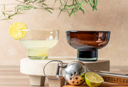 Two handblown glasses with wide rims on pedestal bases, one clear with yellow liquid and lemon slice, one amber-colored with 