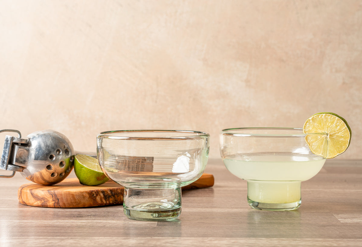 Two clear handblown glasses with wide bowls and pedestal bases, one empty and one filled with pale yellow liquid garnished wi
