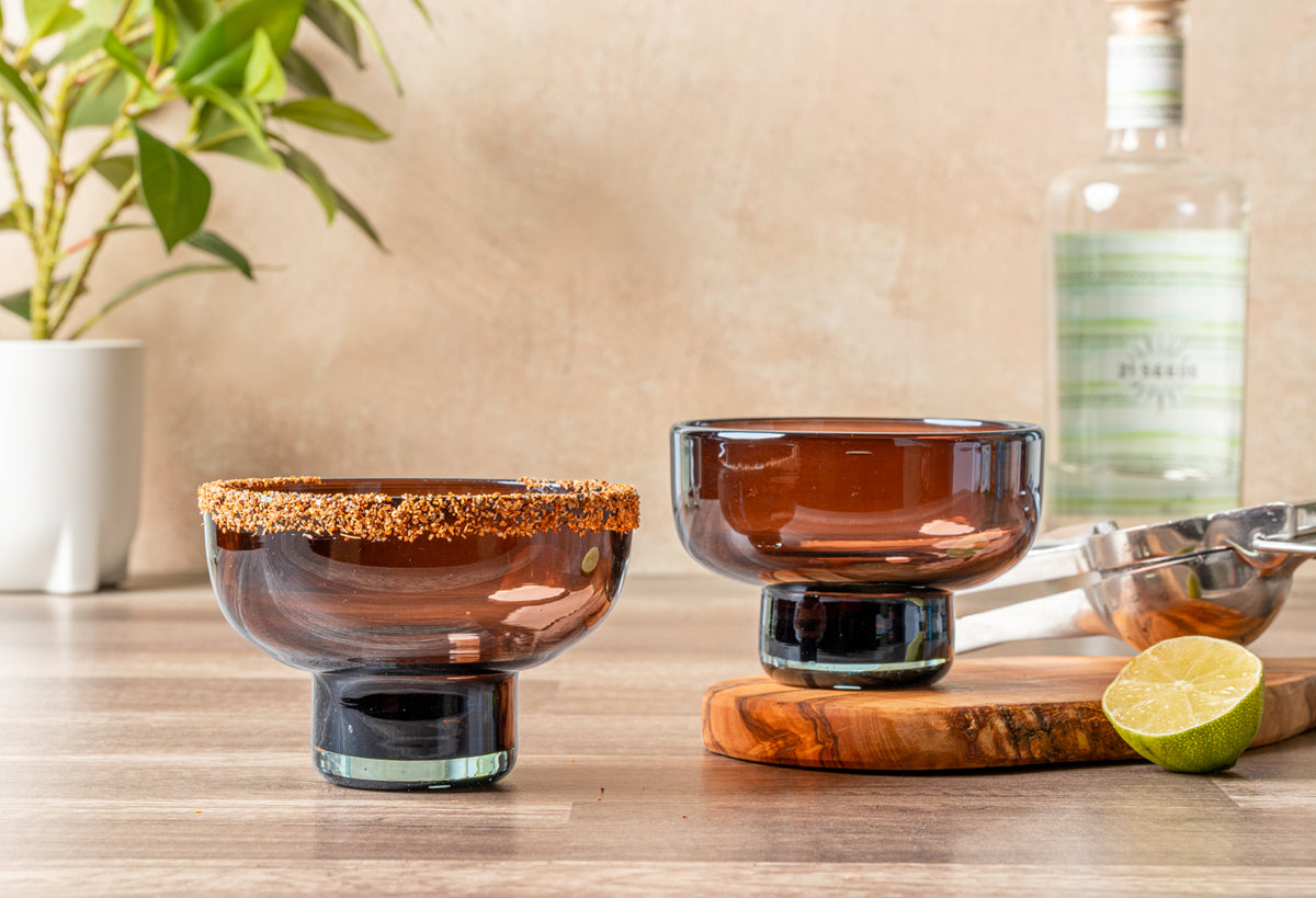 Two handblown amber glasses with wide bowls and pedestal bases, one rimmed with gold sugar, displayed with lime and bar tools