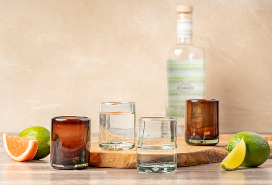 Four handblown shooter glasses in varying amber and clear tones, displayed with citrus and a wooden base.