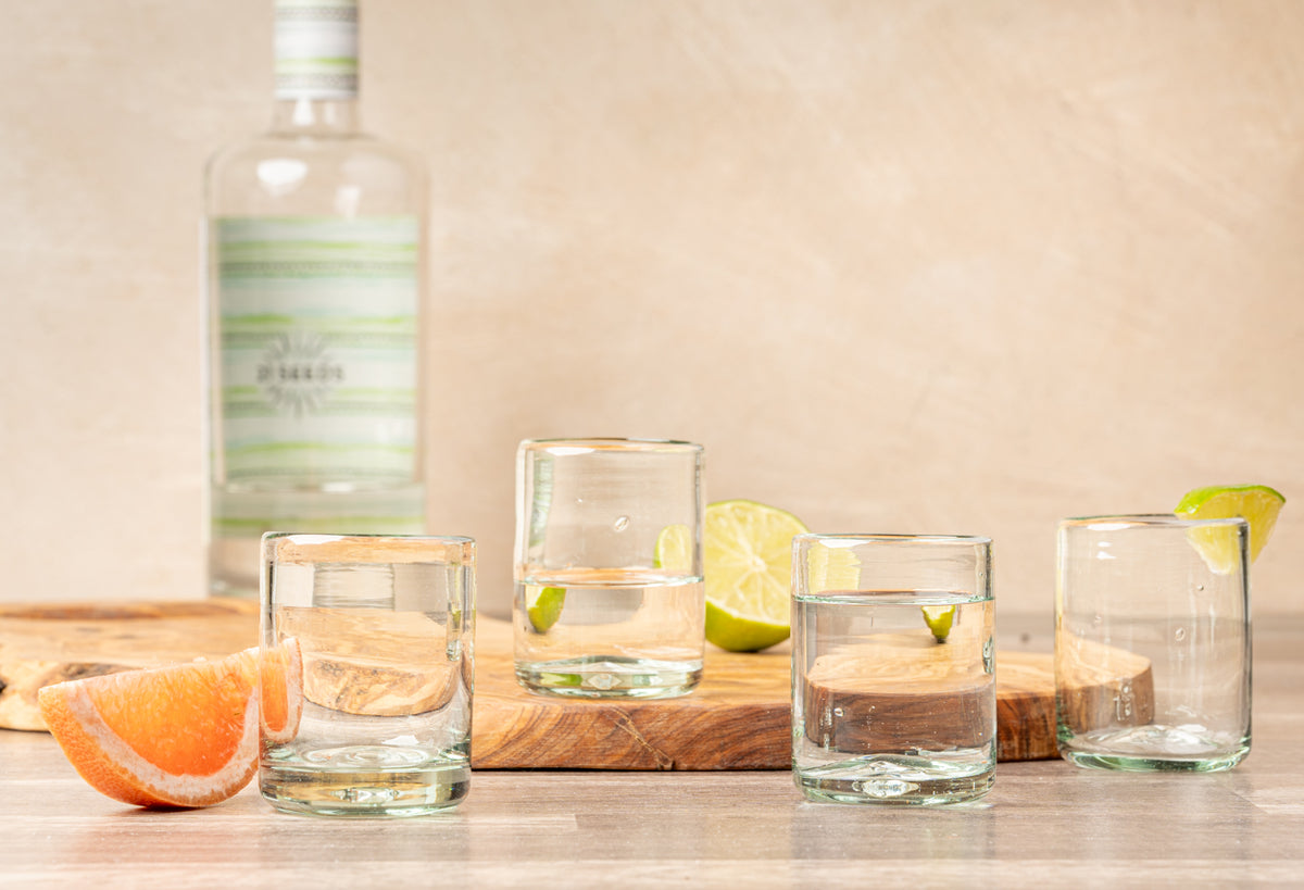 Four clear handblown glasses with gold rims displayed with citrus slices and a bottle on a wooden surface.