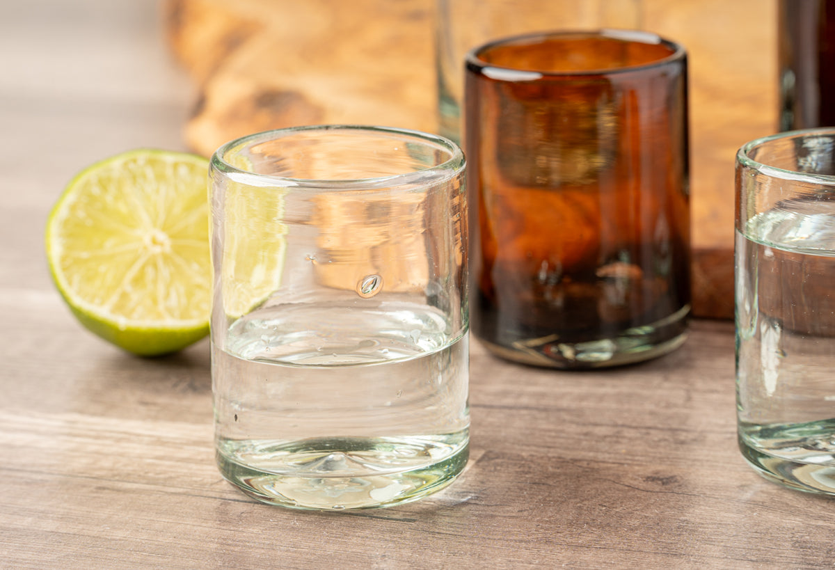 Four handblown shooter glasses in clear and amber glass with a lime wedge on wooden surface.