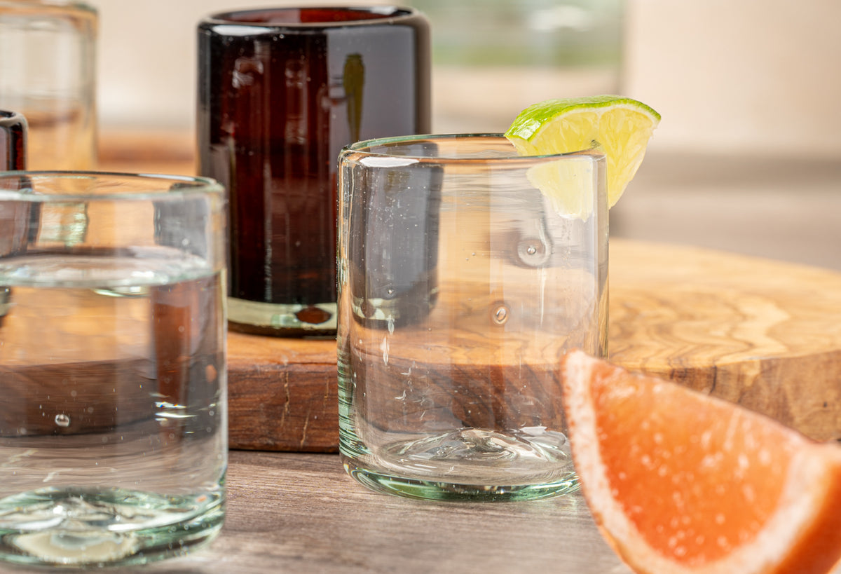 Four clear handblown glasses with varying liquid levels, lime and grapefruit garnishes, on wooden surface with dark bottle.