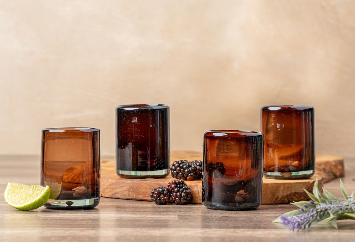 Four handblown shooter glasses with thick bases in varying shades of amber and brown, displayed with lime wedge and berries.