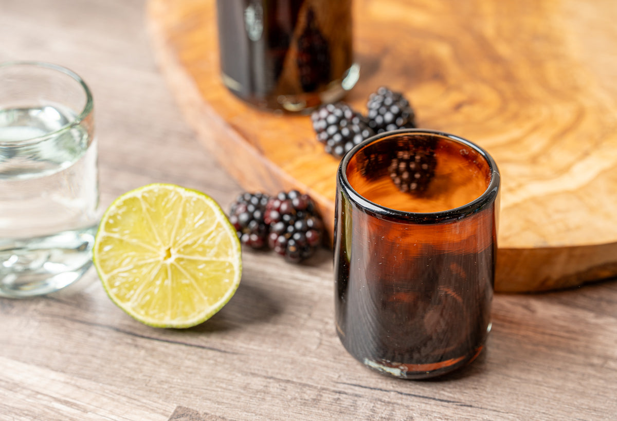 Four small handblown glasses with dark amber liquid, lime slice, and berries on wooden surface.