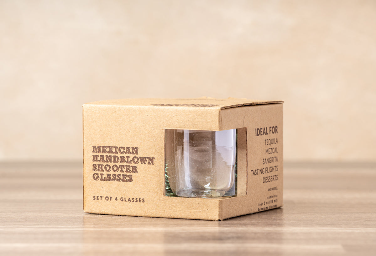 Four clear handblown shooter glasses displayed in kraft cardboard packaging with product text visible.