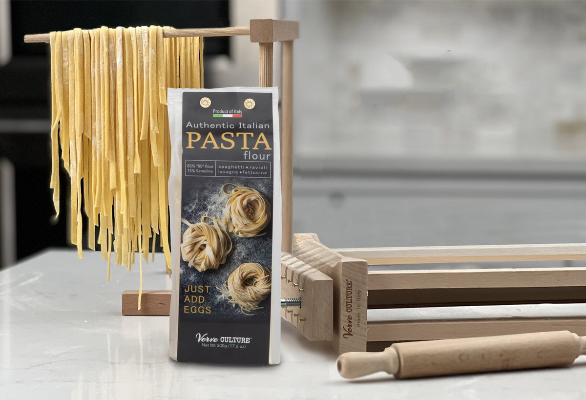 Dried pasta strands hanging on wooden rack next to box of Italian pasta flour and wooden kitchen tools.