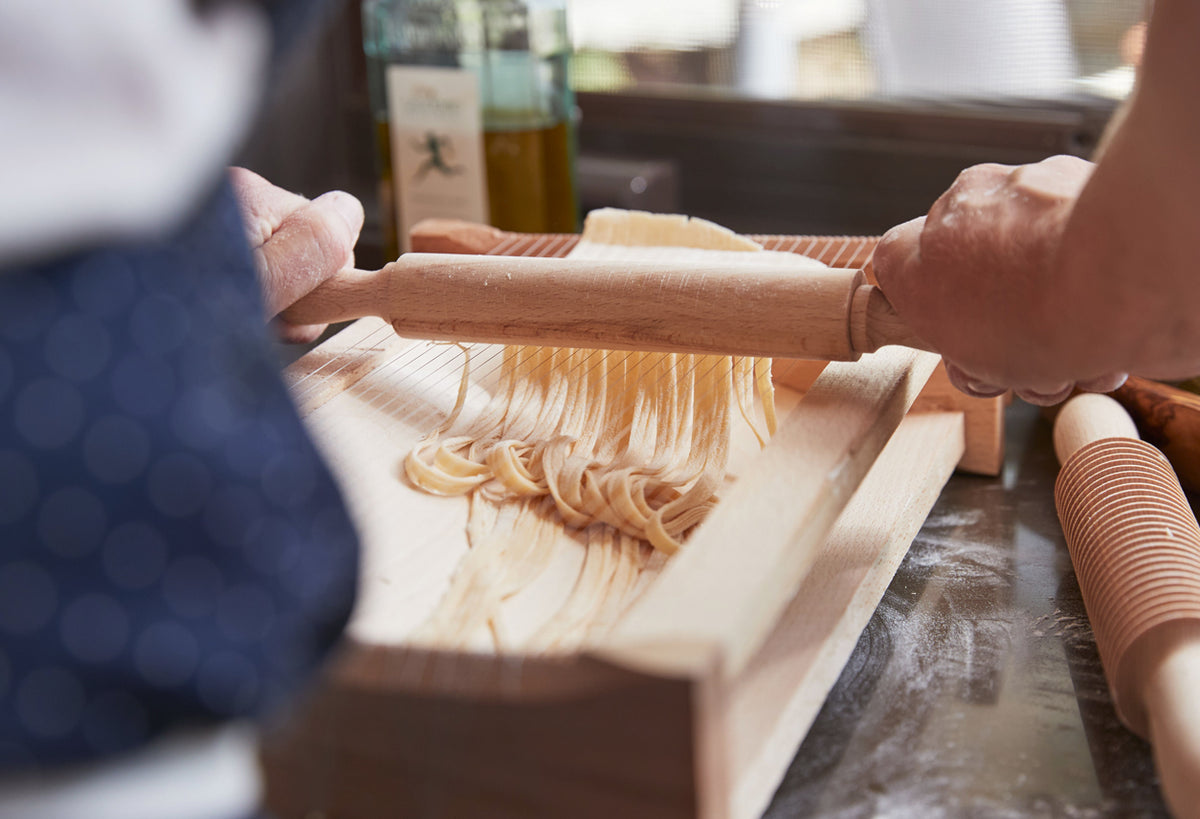 Person rolling fresh pasta with wooden rolling pin over pasta board with oil bottle in background.
