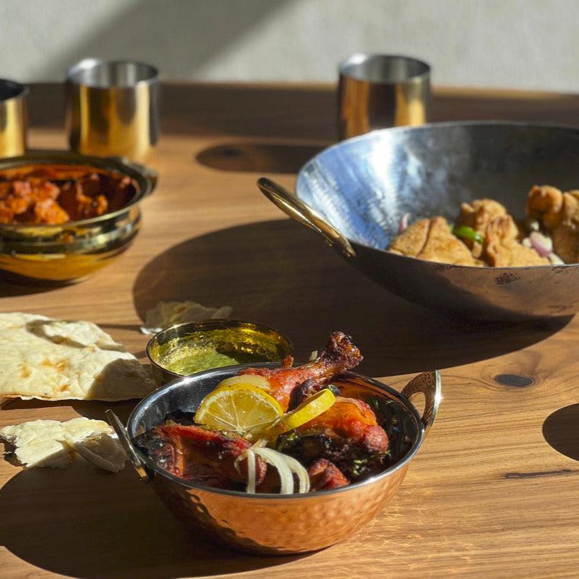 Hammered copper bowls with dark inner lining containing cooked meats and vegetables, displayed on wooden surface with cups an