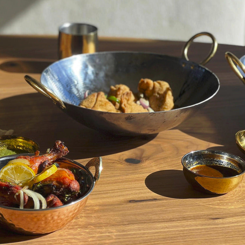Four hammered stainless steel bowls with brass handles containing various foods on a wooden table.