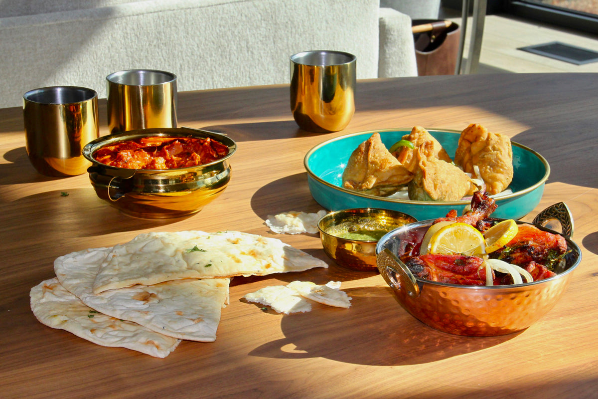 Hammered copper bowls with gold finish displayed with Indian food including curry, bread, and fried snacks on wooden table.