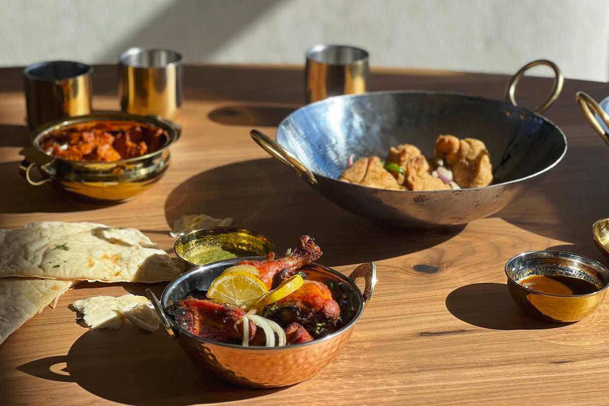 Stainless steel wok with brass handles containing cooked meat and vegetables, displayed with matching brass serveware on wood