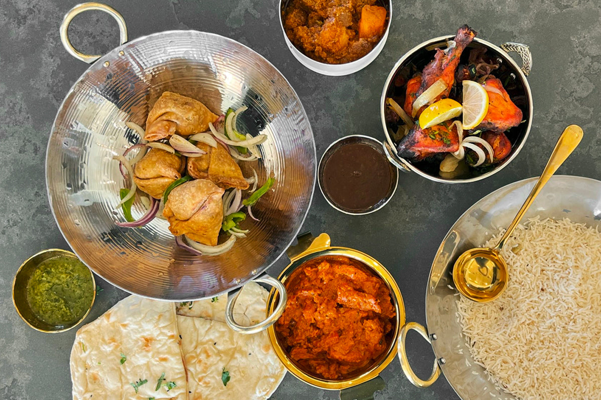 Stainless steel wok with brass handles containing cooked chicken and onions, surrounded by bowls of rice, curry, vegetables, 