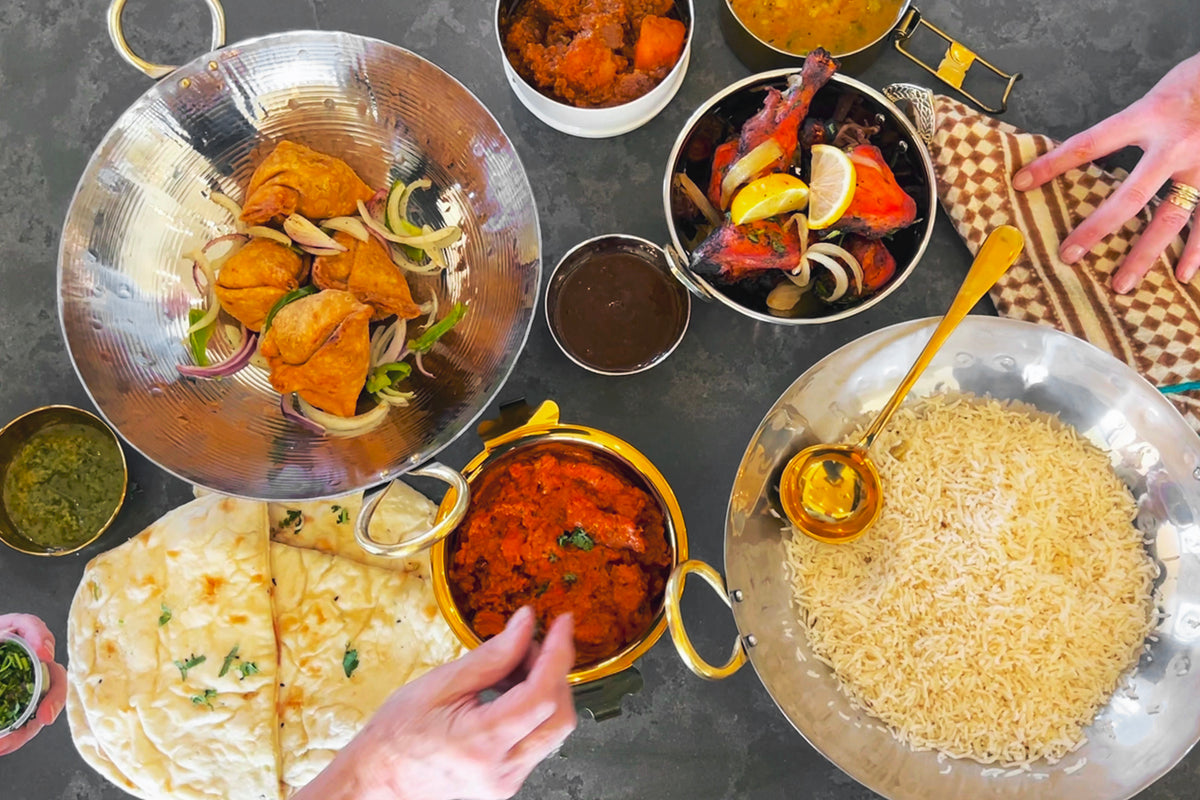 Stainless steel wok with brass handles containing cooked rice, surrounded by bowls of spices and vegetables.