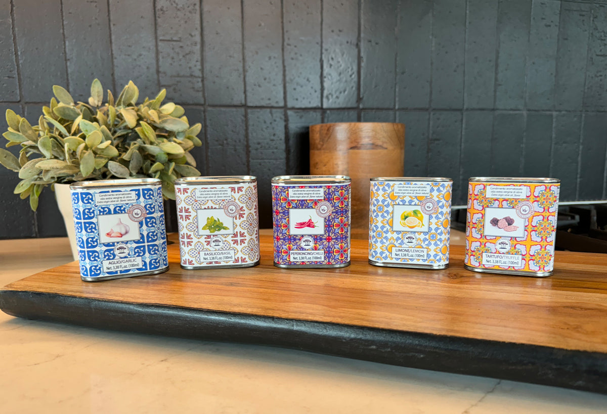 Five decorative tins with patterned labels displayed on wooden surface next to potted plant and wooden container.