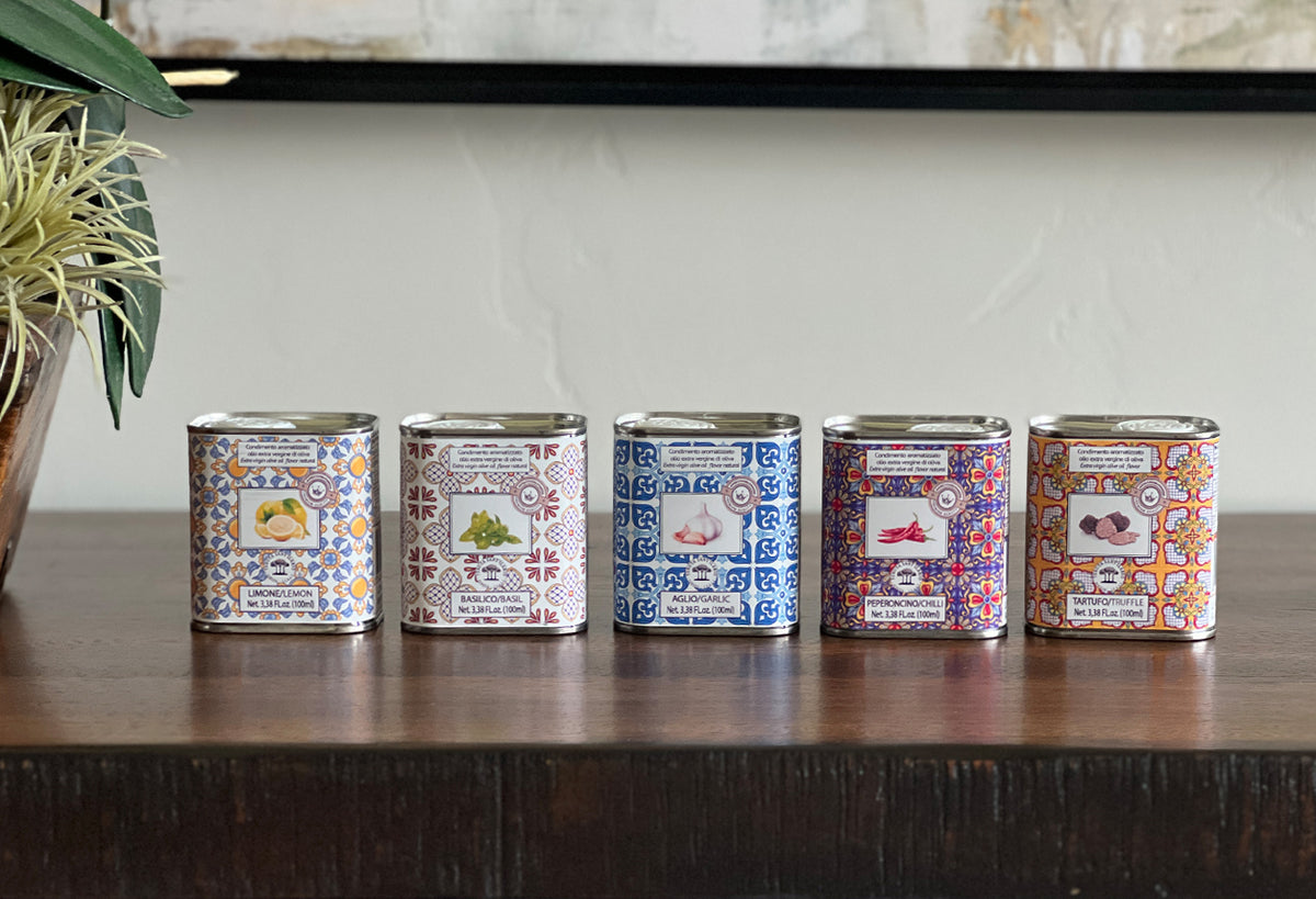 Five decorative tins with patterned labels in blue, orange, and red designs containing olive oil variants.