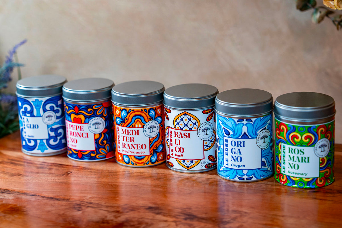 Six decorative metal tins with colorful Italian patterns containing dried herbs and spices.