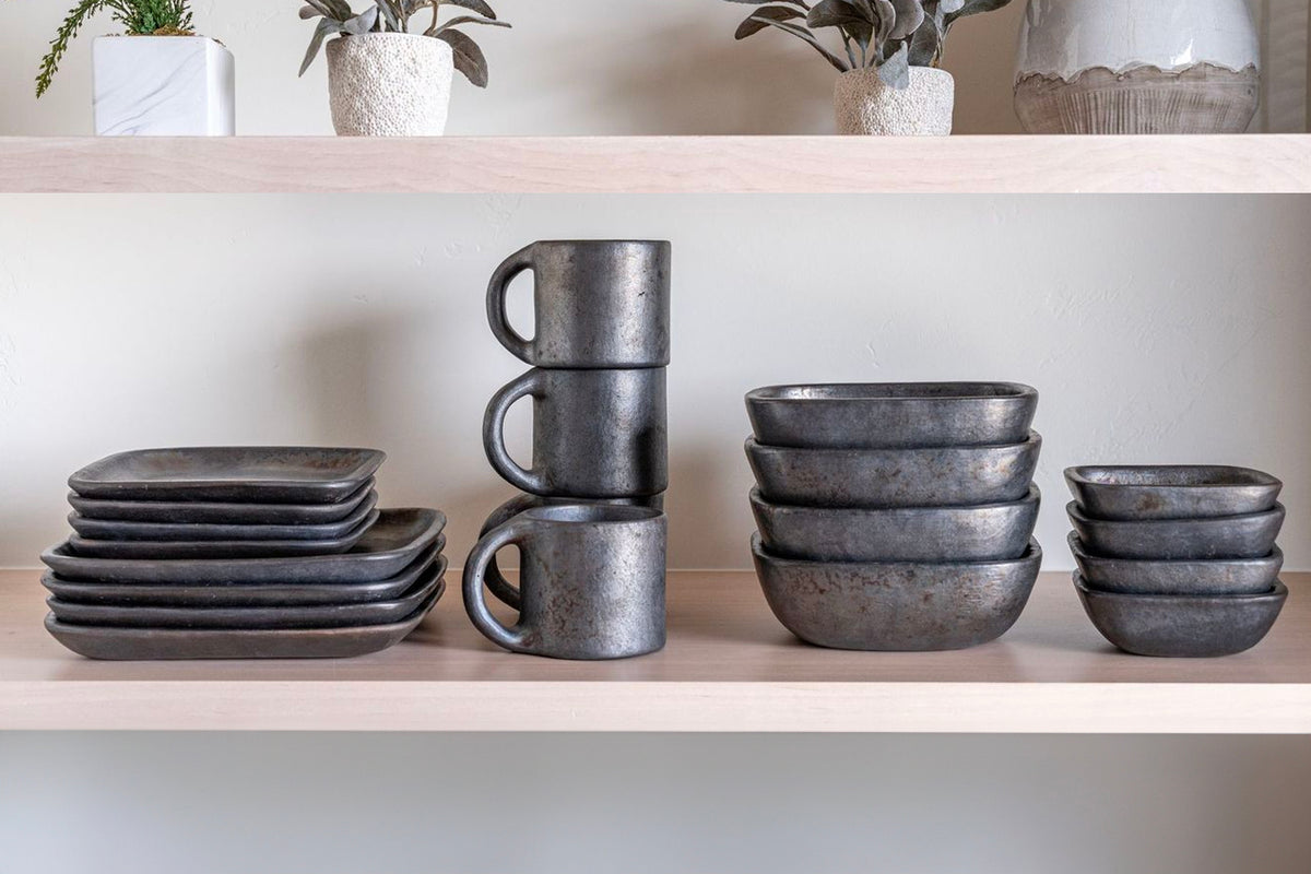 Stacked dark gray pottery plates, bowls, and mugs with textured matte finish on wooden shelf.