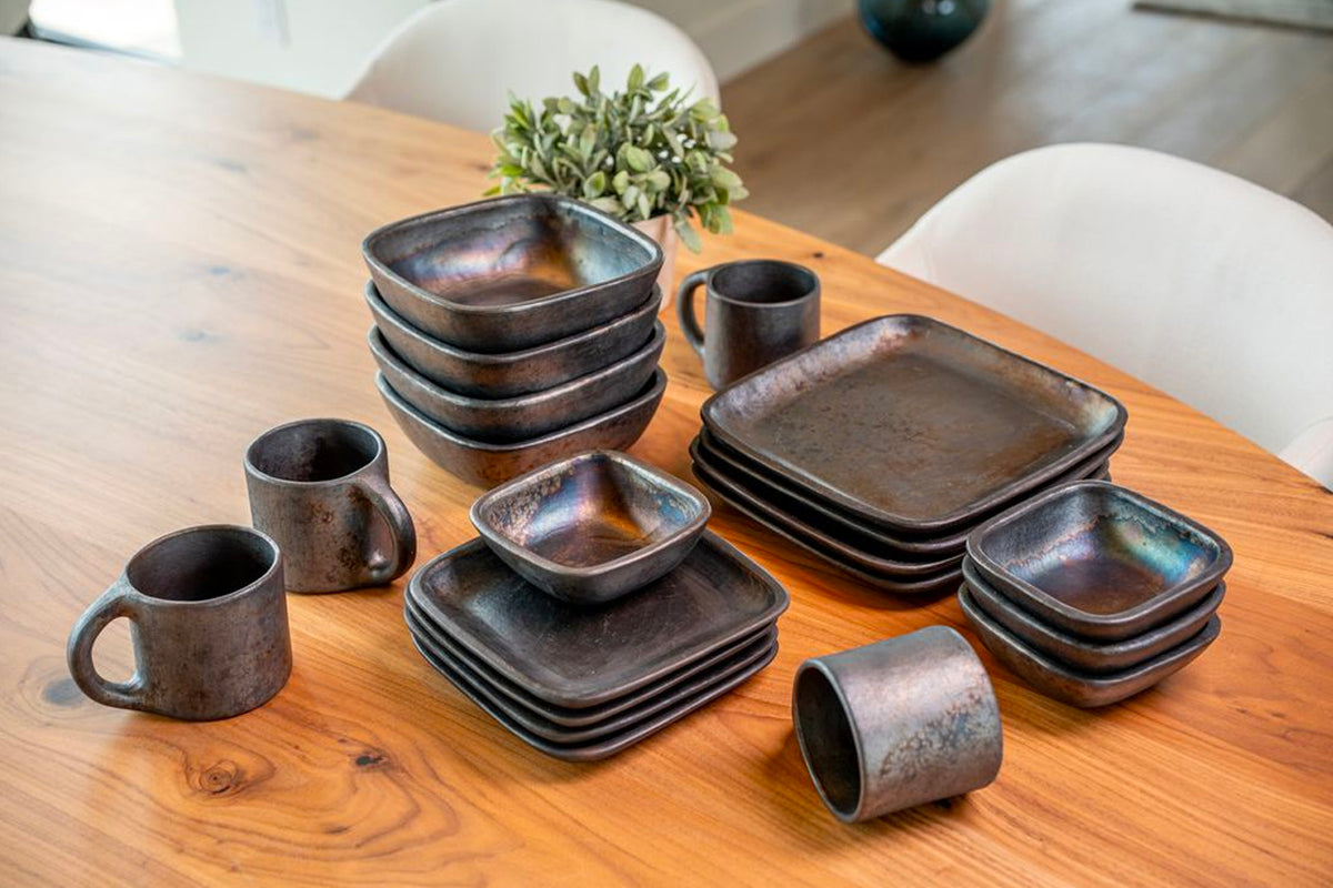 Dark brown square pottery dinnerware set with mugs and bowls arranged on wooden table.