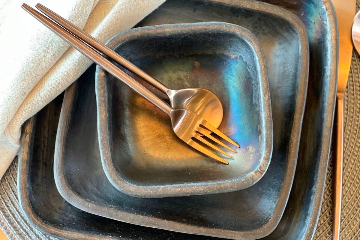 Stacked dark gray pottery plates with rounded edges, fork, and cream napkin on woven surface.