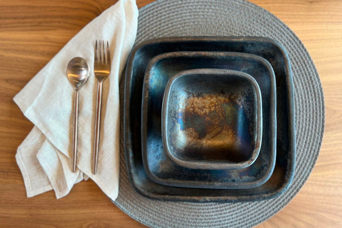 Three nesting handmade pottery bowls with dark glaze and textured surfaces on a woven placemat.