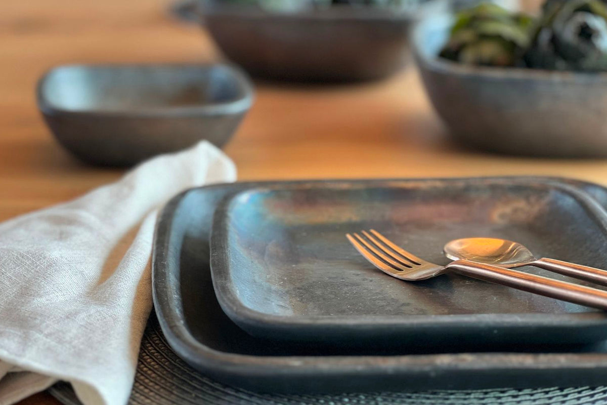 Dark gray pottery dinner plate with a fork and spoon on it, styled on a table with napkin and bowls.