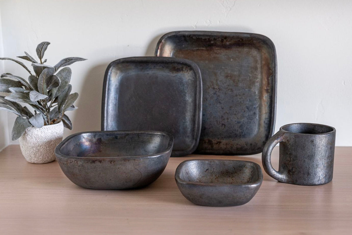 Black pottery dinnerware set including large square plate, bowls, mug, and small dish with textured finish.