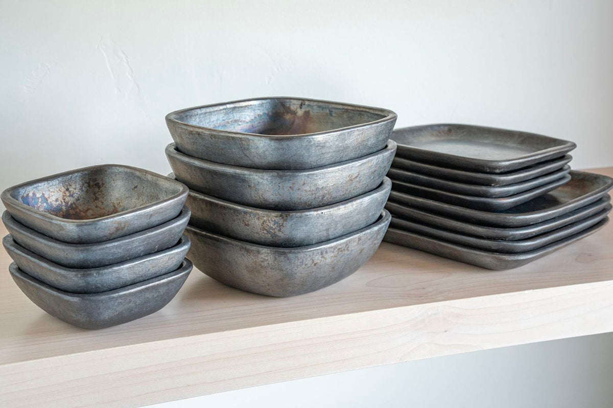 Gray square pottery bowls stacked in groups on a wooden shelf, displaying various sizes and rounded corners.