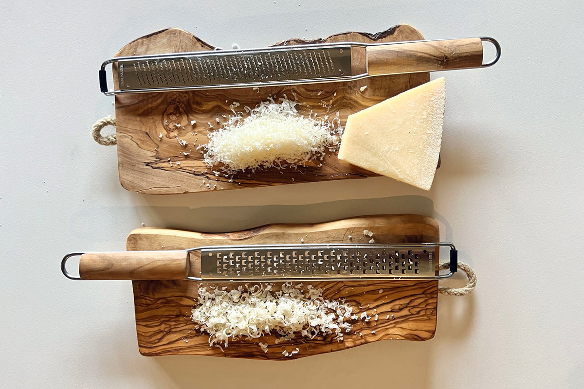 Two olivewood paddle graters with metal grating surfaces and wooden handles, each with shredded cheese on its board.