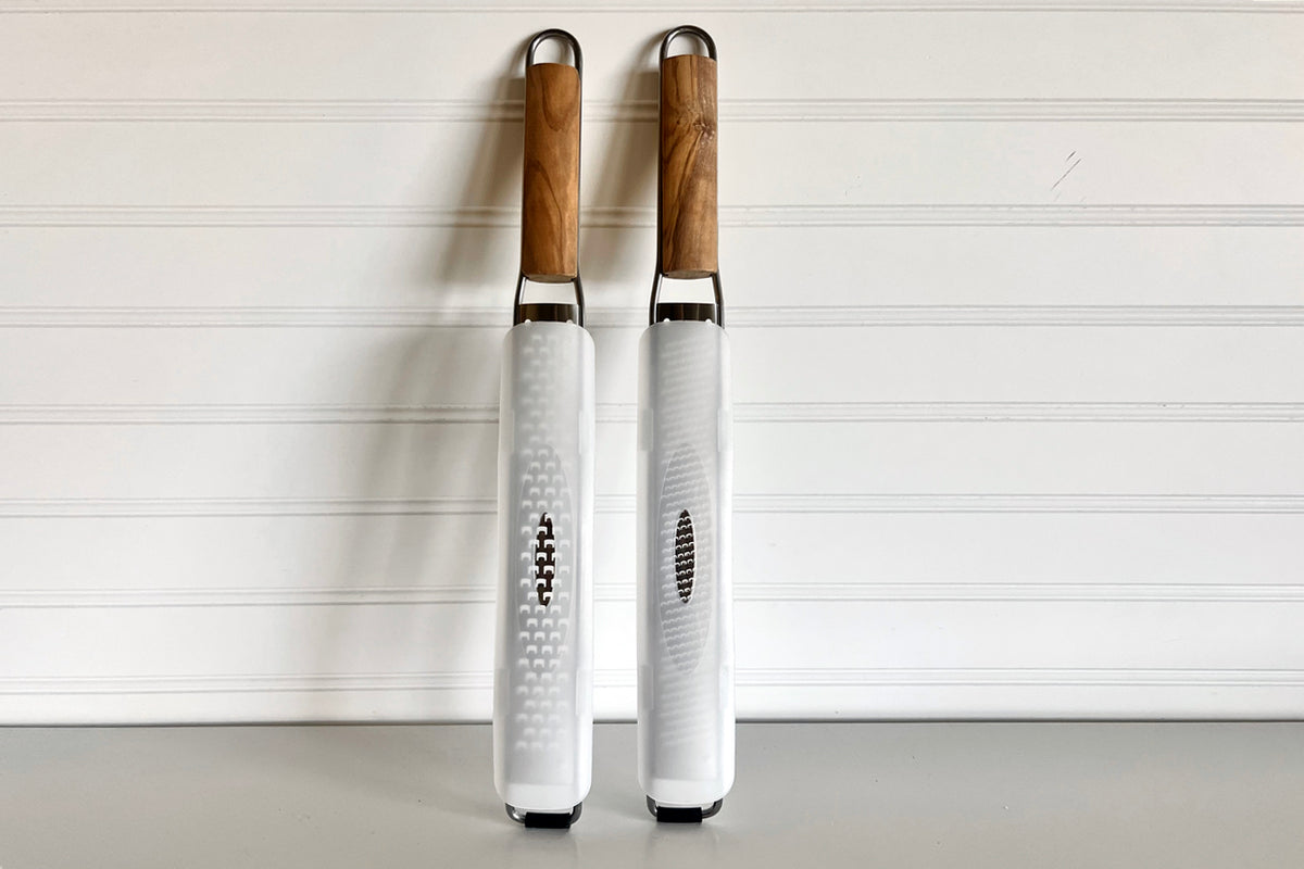 Two paddle graters with olive wood handles and stainless steel grating surfaces, one with fine holes and one with larger hole