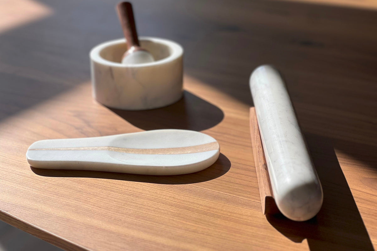 White marble mortar with brown wooden pestle, marble spoon, and rolling pin arranged on wooden surface.