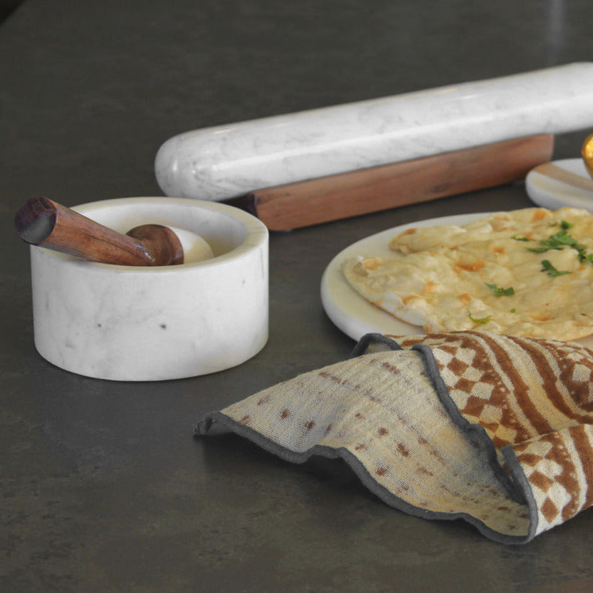 White marble mortar and pestle with wooden handle, displayed with a matching pestle and kitchen items on dark surface.