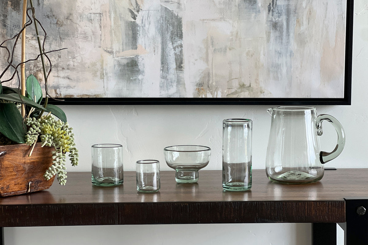 Four clear handblown glass shooter glasses with varied shapes displayed on a wooden shelf.