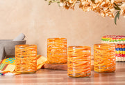 Four orange handblown glasses with white swirl patterns displayed on a wooden surface with decorative items.