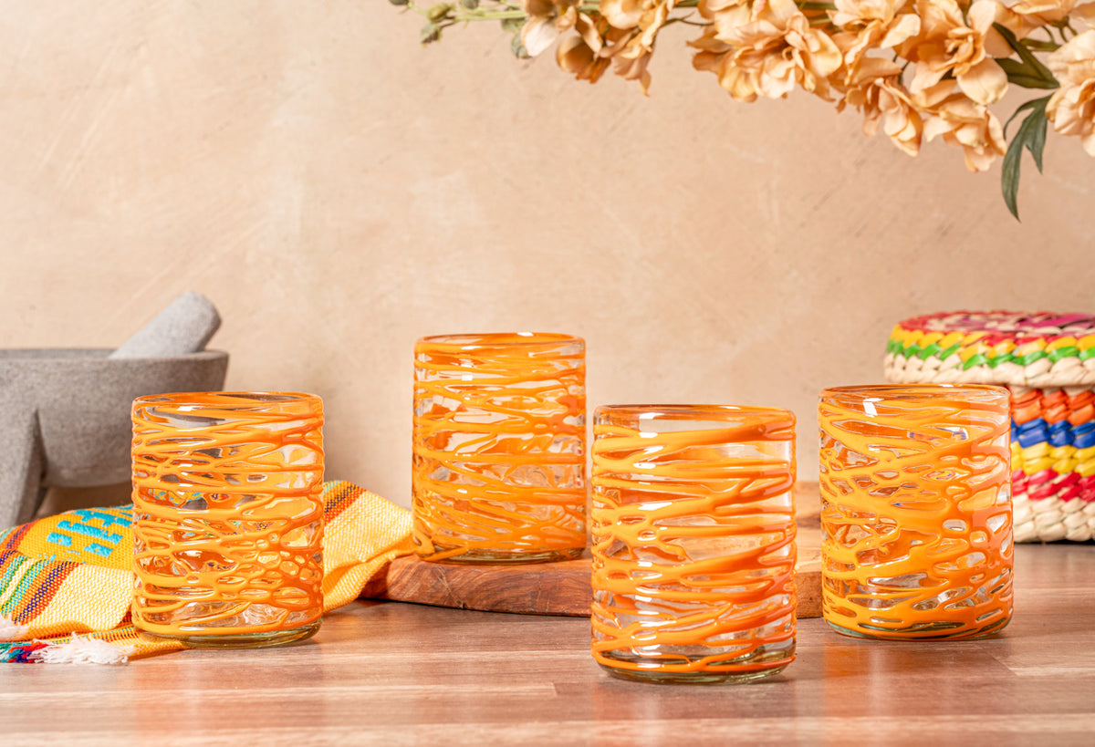 Four orange handblown glasses with white swirl patterns displayed on a wooden surface with decorative items.