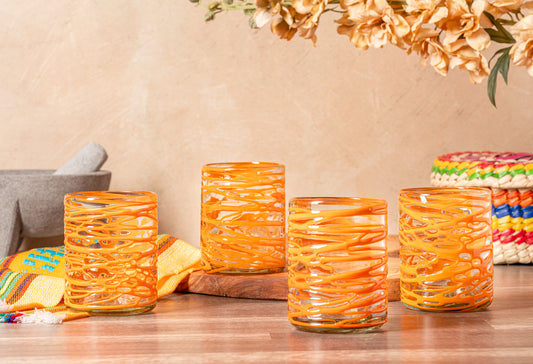 Four orange handblown glasses with white swirl patterns displayed on a wooden surface with decorative items.