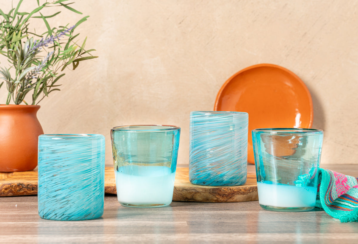 Four handblown glasses with turquoise swirl patterns and frosted bases arranged on wood with orange ceramic bowl and plant.