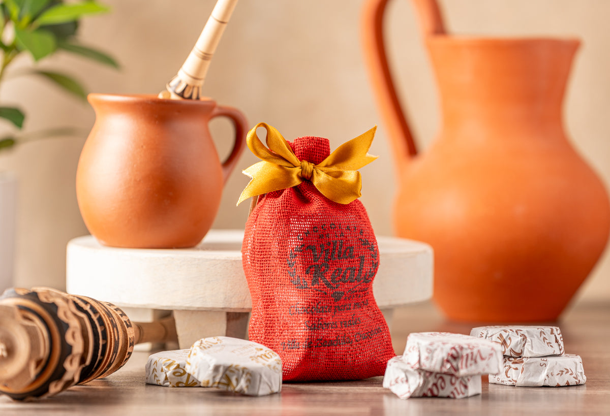 Red fabric pouch with gold bow surrounded by individual wrapped chocolate portions and terracotta serving vessels with wooden