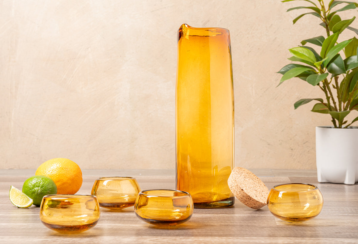 Tall amber glass bottle with golden liquid surrounded by four matching tasting glasses, citrus fruits, cork, and potted plant