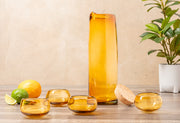Tall amber glass bottle with golden liquid surrounded by four matching tasting glasses, citrus fruits, cork, and potted plant