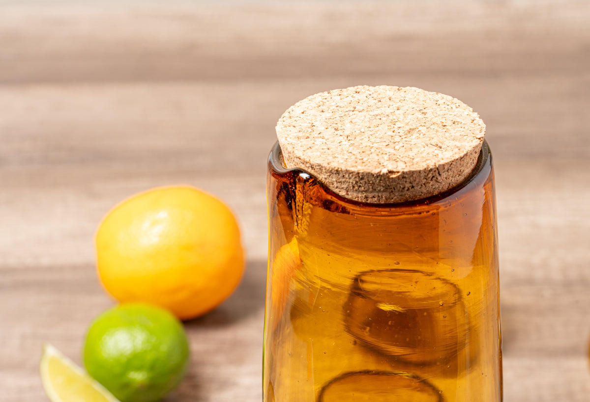 Glass jar with cork stopper filled with amber liquid and ice, surrounded by yellow lemon and green lime on wooden surface.