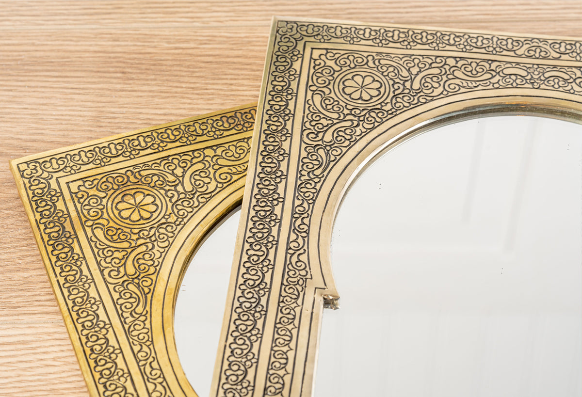 Brass-framed mirror with ornate geometric and floral engraved patterns on cream background, displayed on wooden surface.