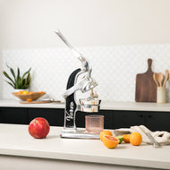 Manual citrus juicer with black and silver lever design on white countertop with citrus fruits and glass.