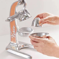 Manual citrus juicer with metal frame, orange enamel panel, lever press, and removable juice cups being held by hands.