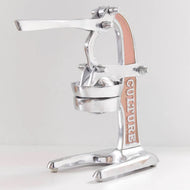 Manual citrus juicer with polished metal frame, pink grip accents, lever arm, and collection bowl on stable base.